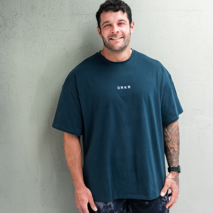 Smiling man wearing the forest green Endure Oversized Tee against a white wall
