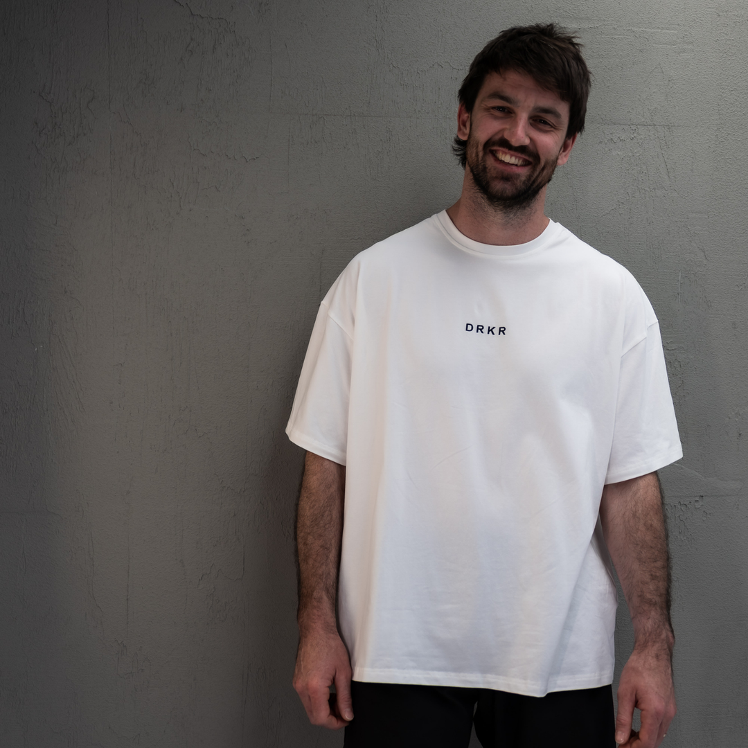 Smiling man wearing the white Endure Oversized Tee against a concrete wall
