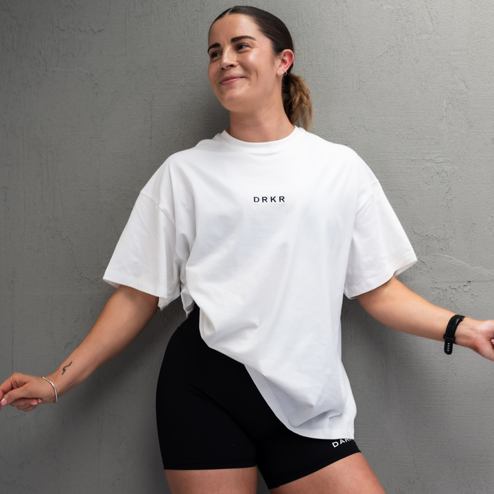Smiling woman wearing the white Endure Oversized Tee with black shorts against a concrete wall