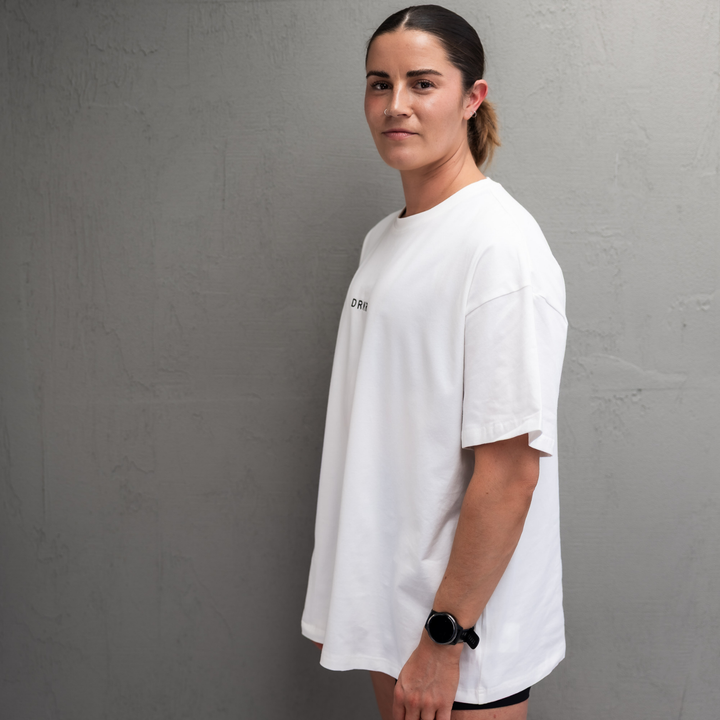 Woman wearing the white Endure Oversized Tee against a concrete wall, side view