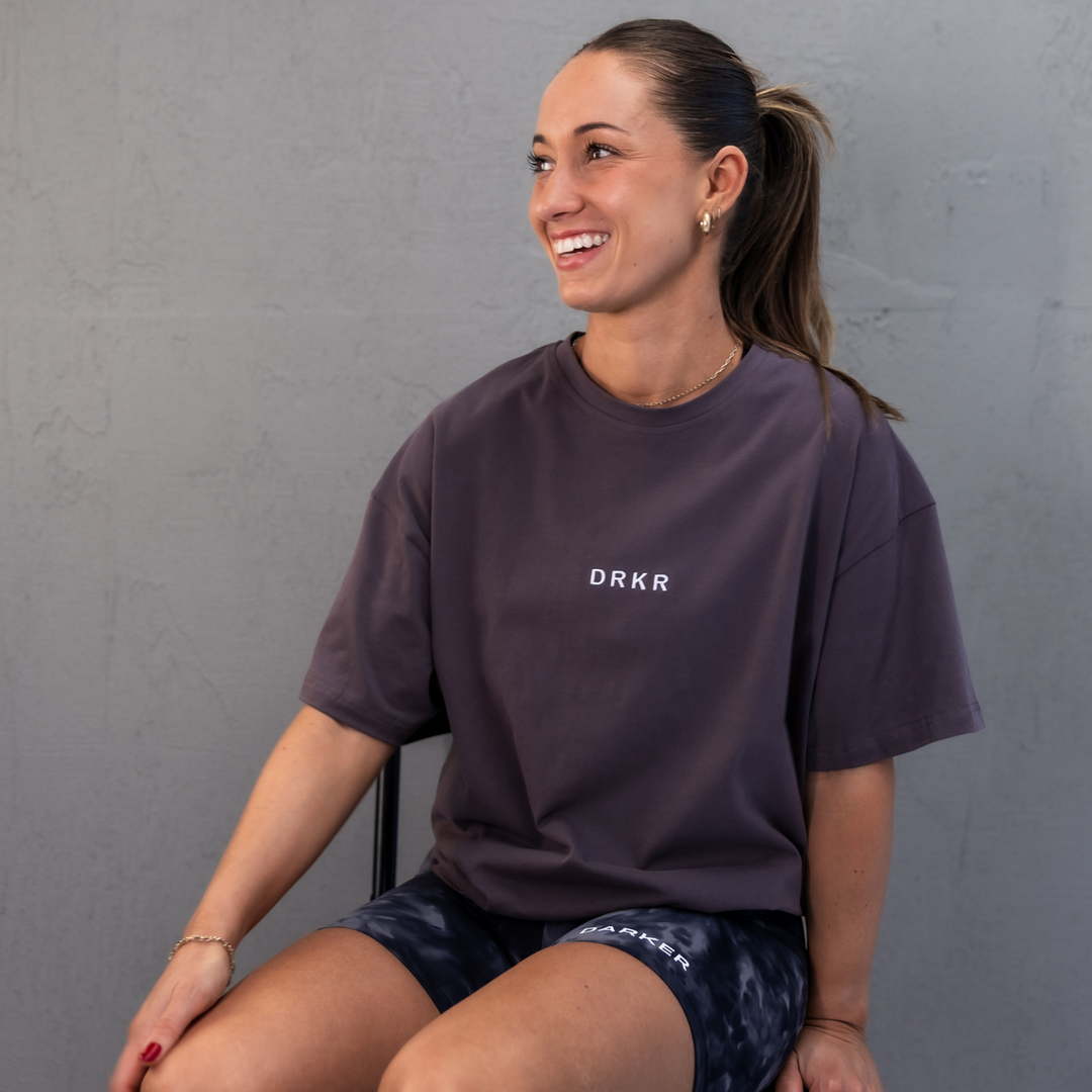 Smiling woman seated in the storm grey Endure Oversized Tee against a concrete wall