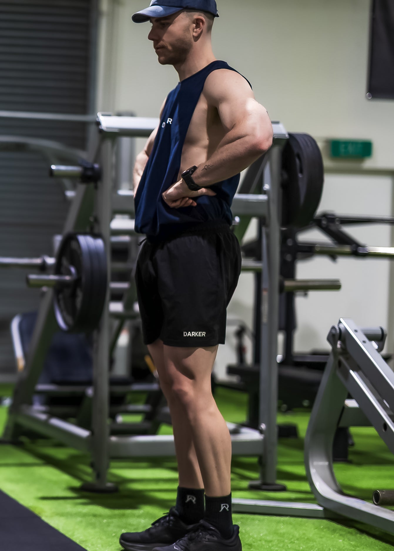 Muscular man in gym wearing a cap and black shorts with visible branding.