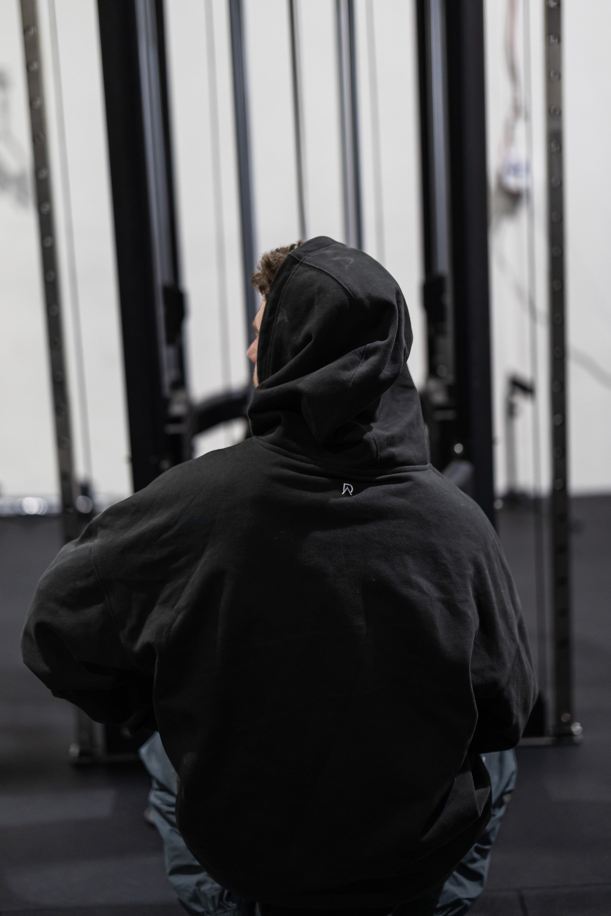 Person wearing a black hoodie with a logo, sitting in a gym.