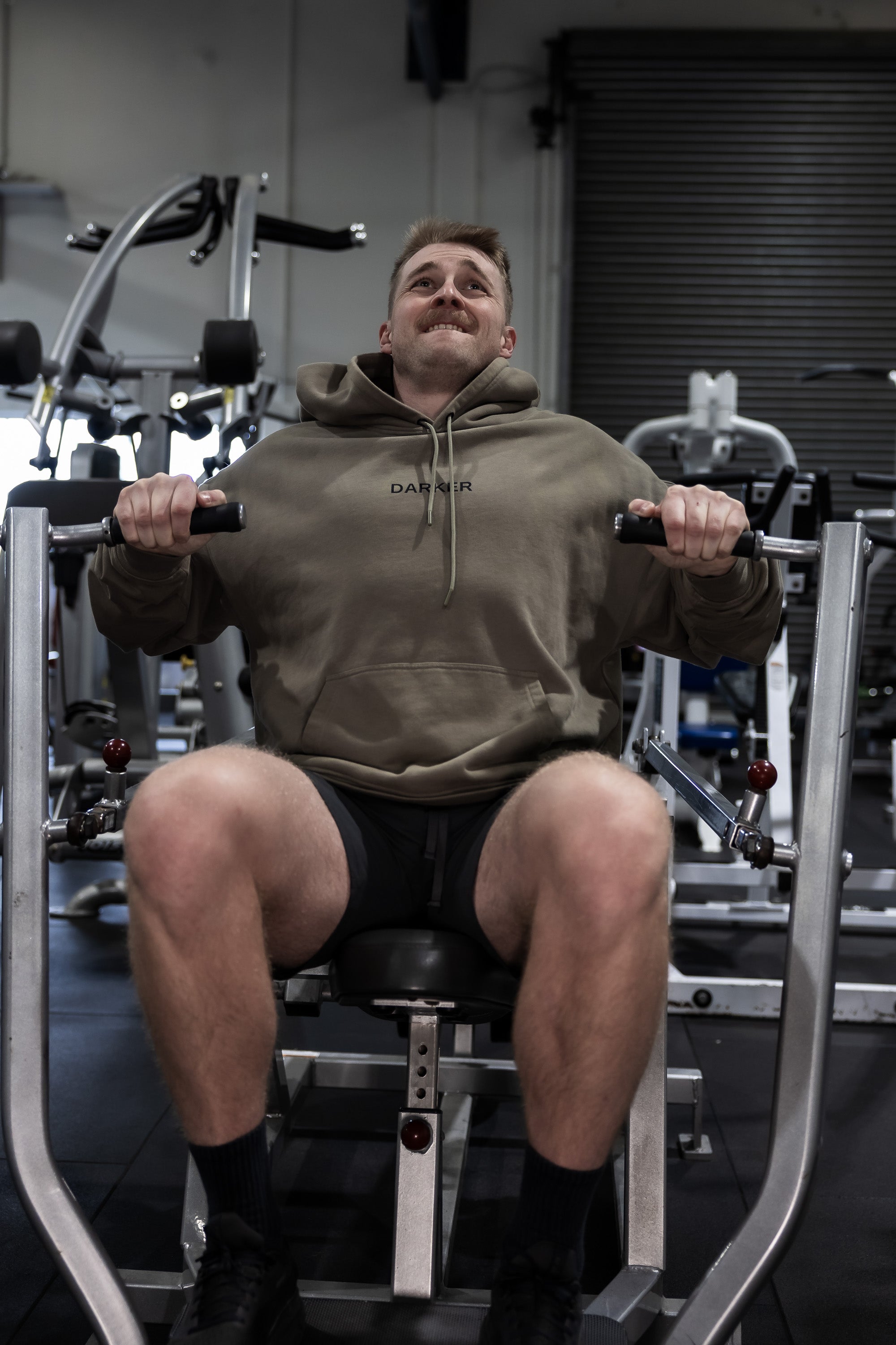 Man exercising on a weight machine in a gym wearing a khaki hoodie with branding.