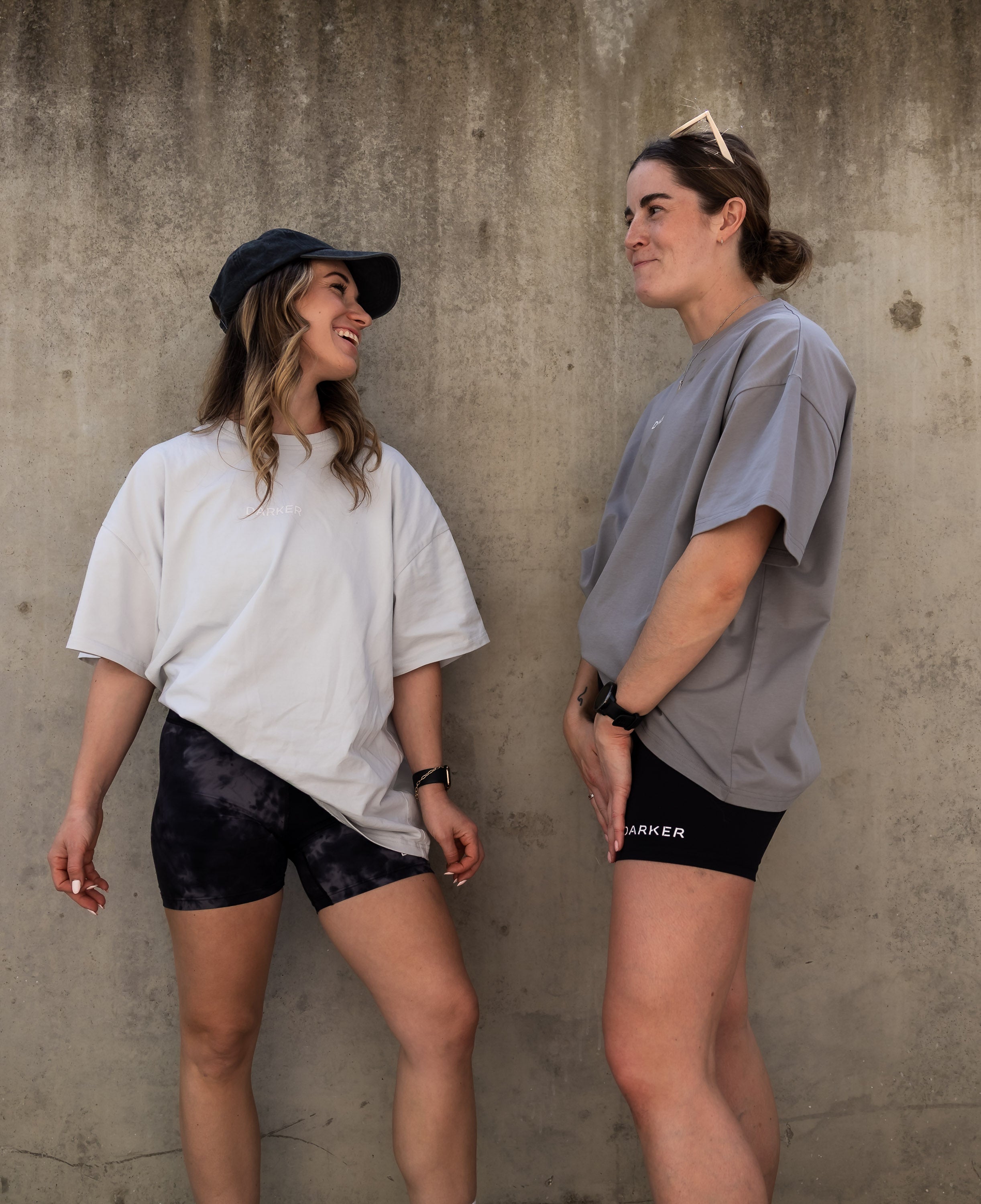 Two women standing against a concrete wall, wearing casual outfits.