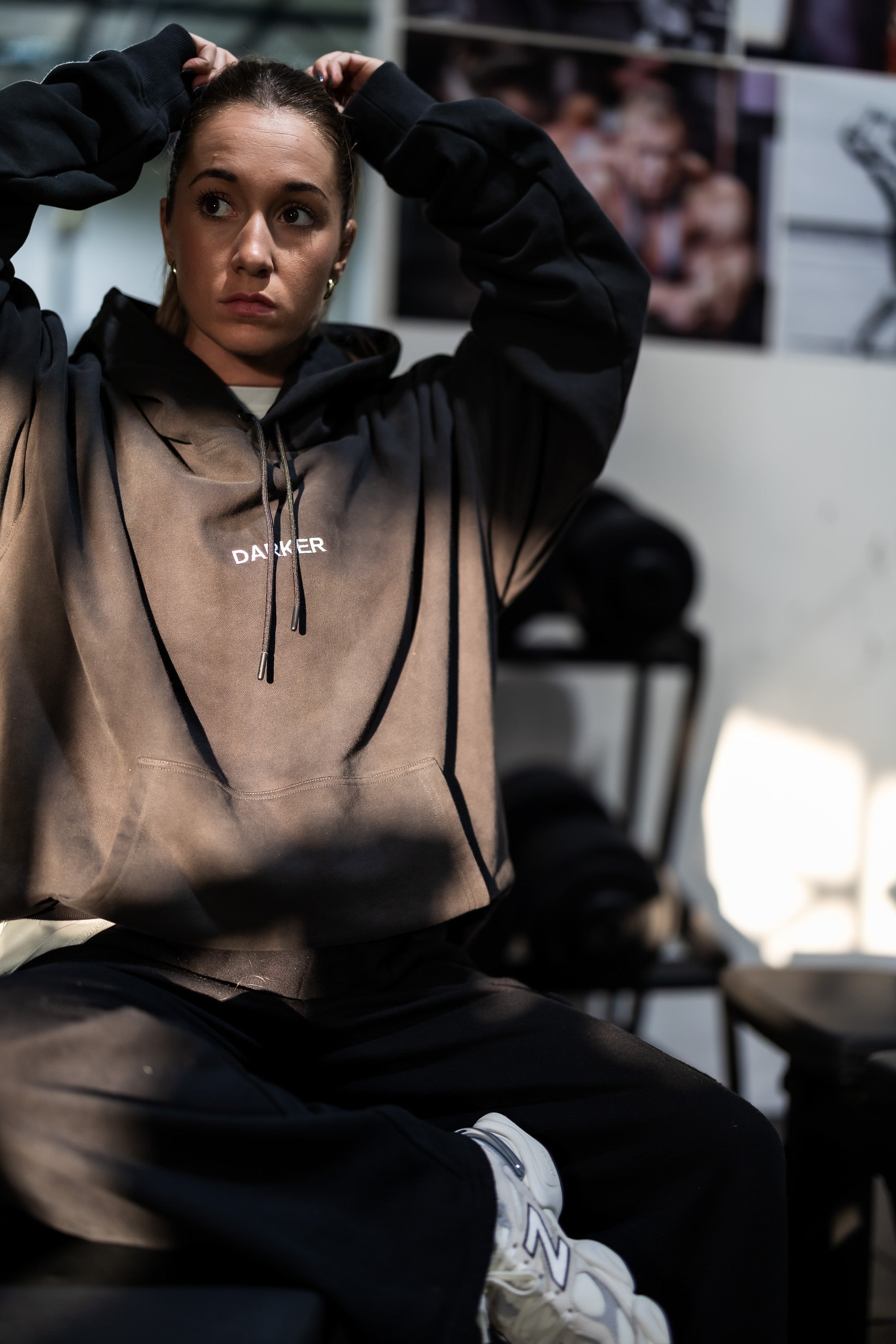 Person wearing a black hoodie with a darker logo, sitting in a room with posters on the wall.