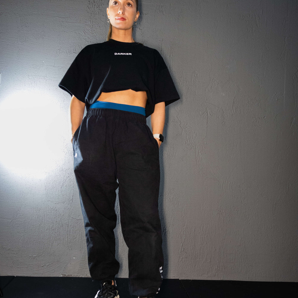 Women in black baggy drift pants wearing a black tucked in t-shirt with DARKER on the chest. Is wearing blue shorts underneath