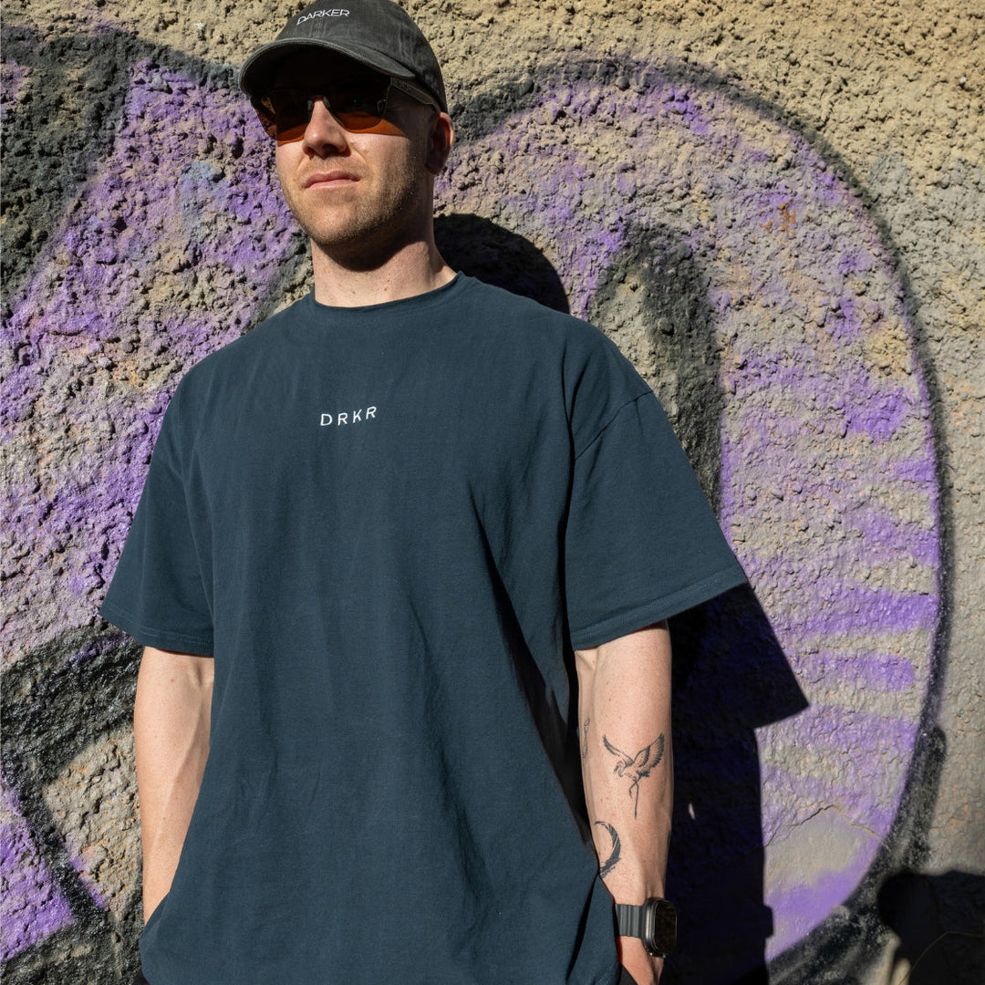 Man in a cap wearing the forest green Endure Oversized Tee against a graffiti wall