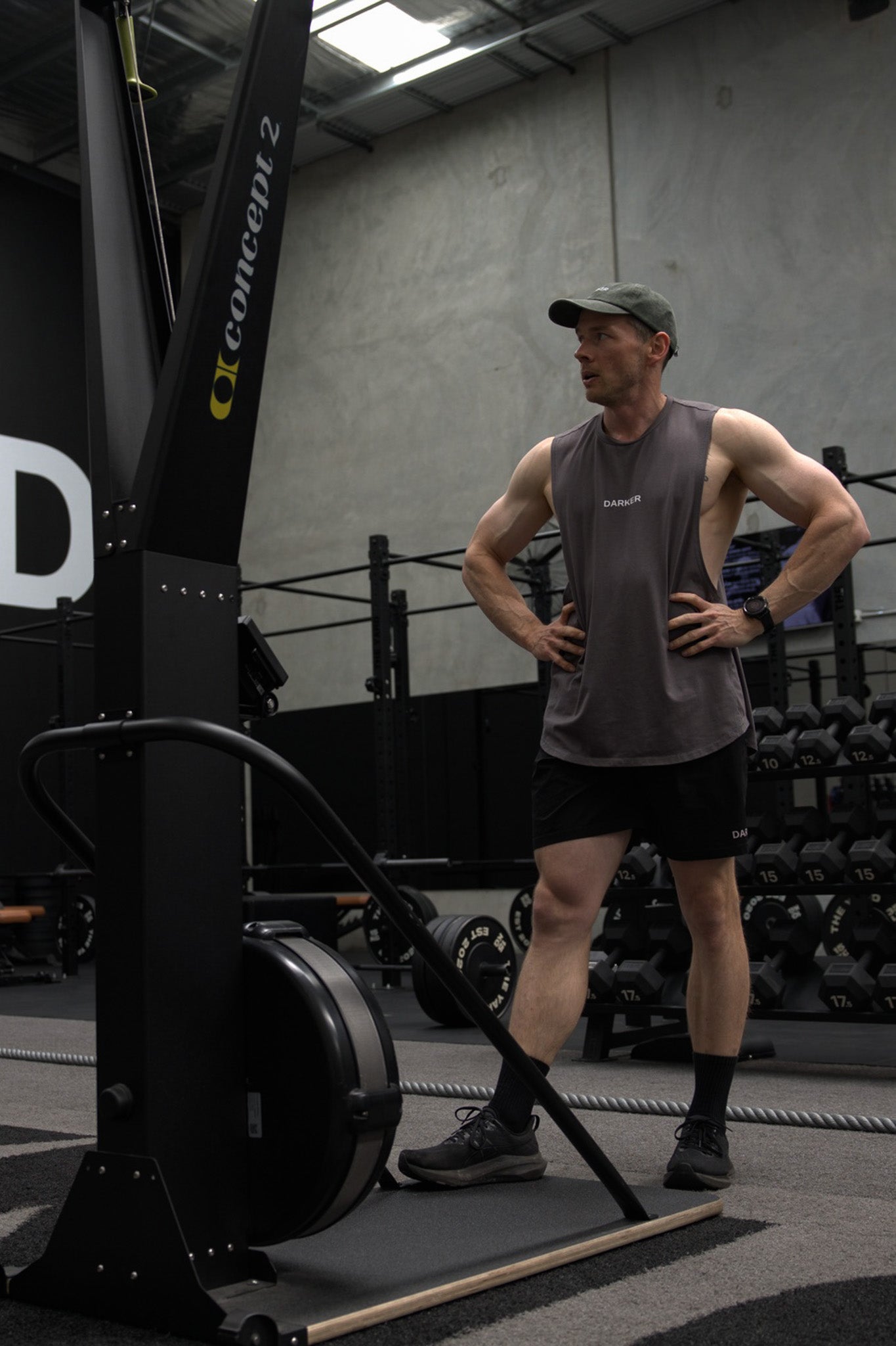A man in a gym stands next to a Concept 2 rowing machine, wearing a gray sleeveless shirt, black shorts, and a green cap.
