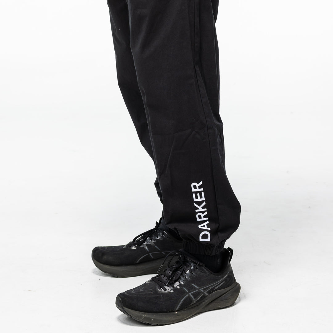 Black pants with 'DARKER' branding worn by a person on a white background. Shot close-up from the knee down to the shoes