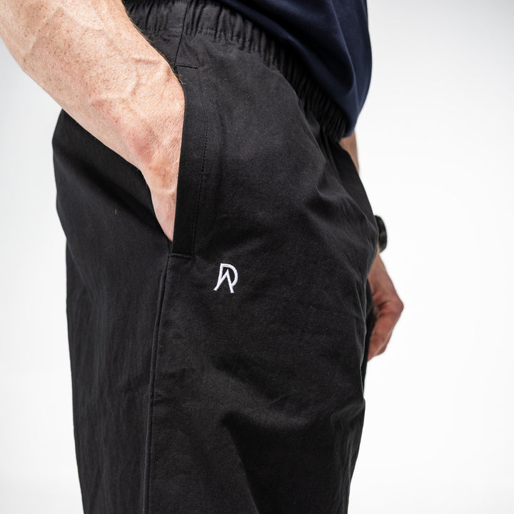 Black pants with a white logo on a plain background where the model has his hands in the pocket