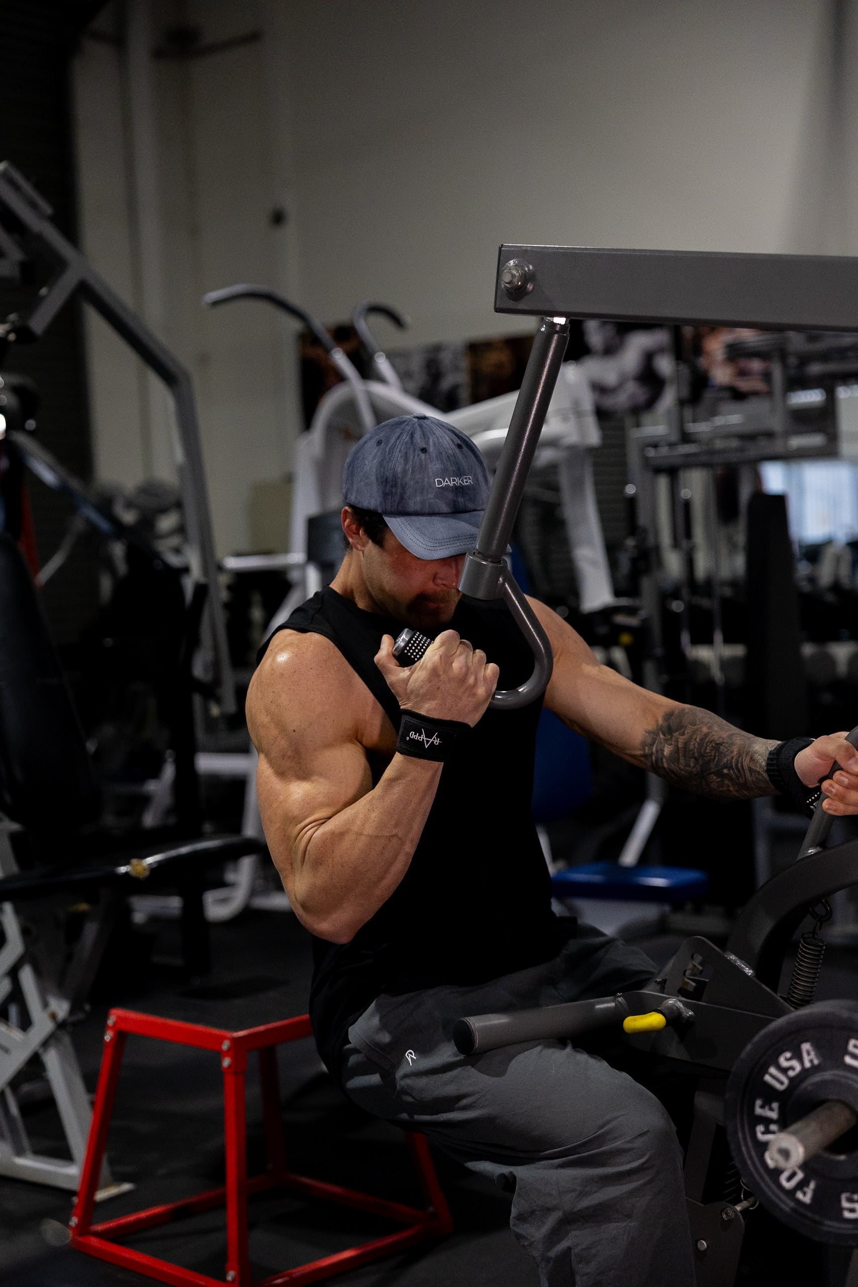 A man in a gym wearing a black tank top, gray sweatpants, and a blue cap, performing a seated arm exercise on a machine. Gym equipment is visible in the background.