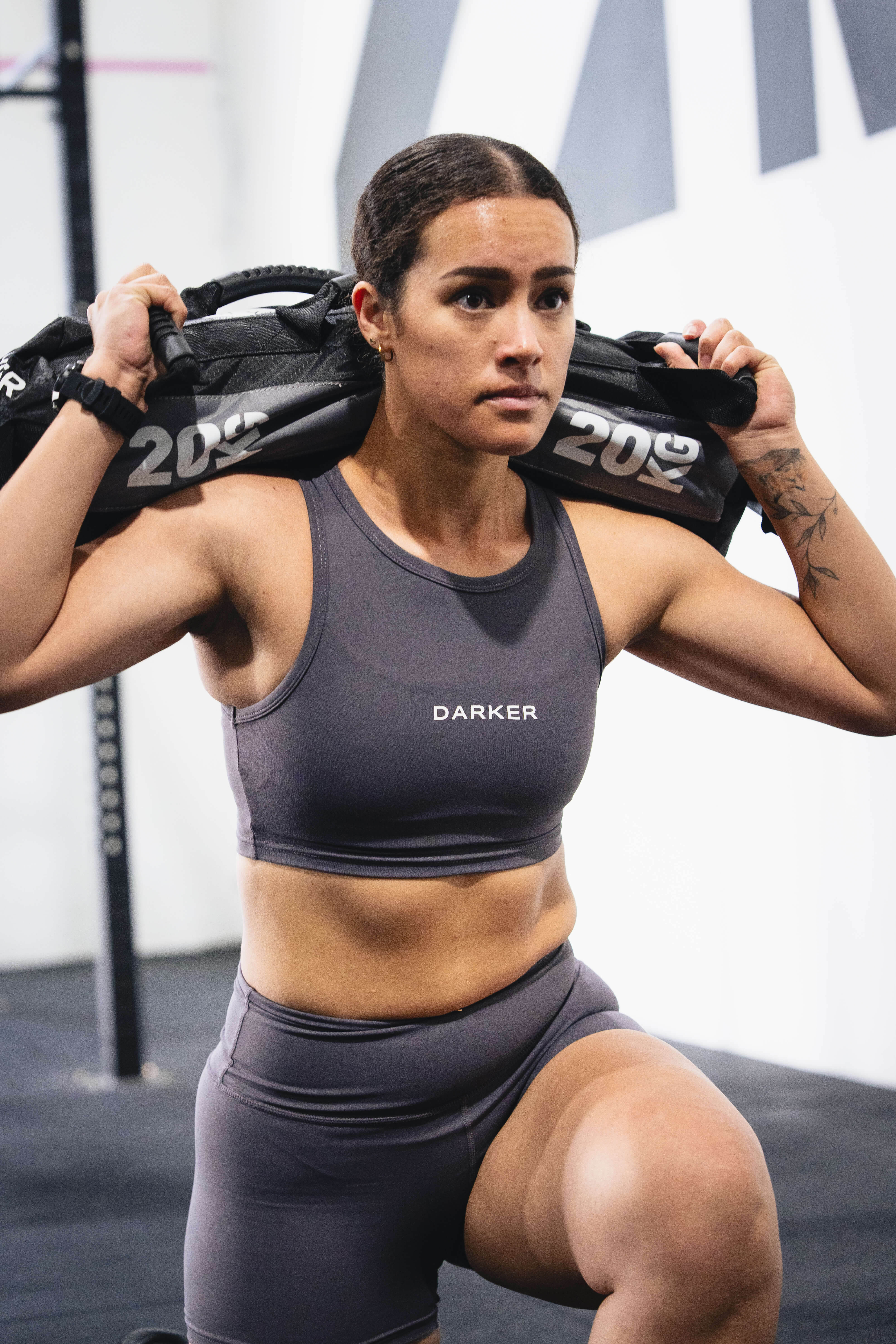 A woman in a grey agile cropped tank and matching shorts holds a 20 kg weight sand bag across her shoulders in a gym setting. The brand name 'DARKER' is printed on the front of her cropped tank. 