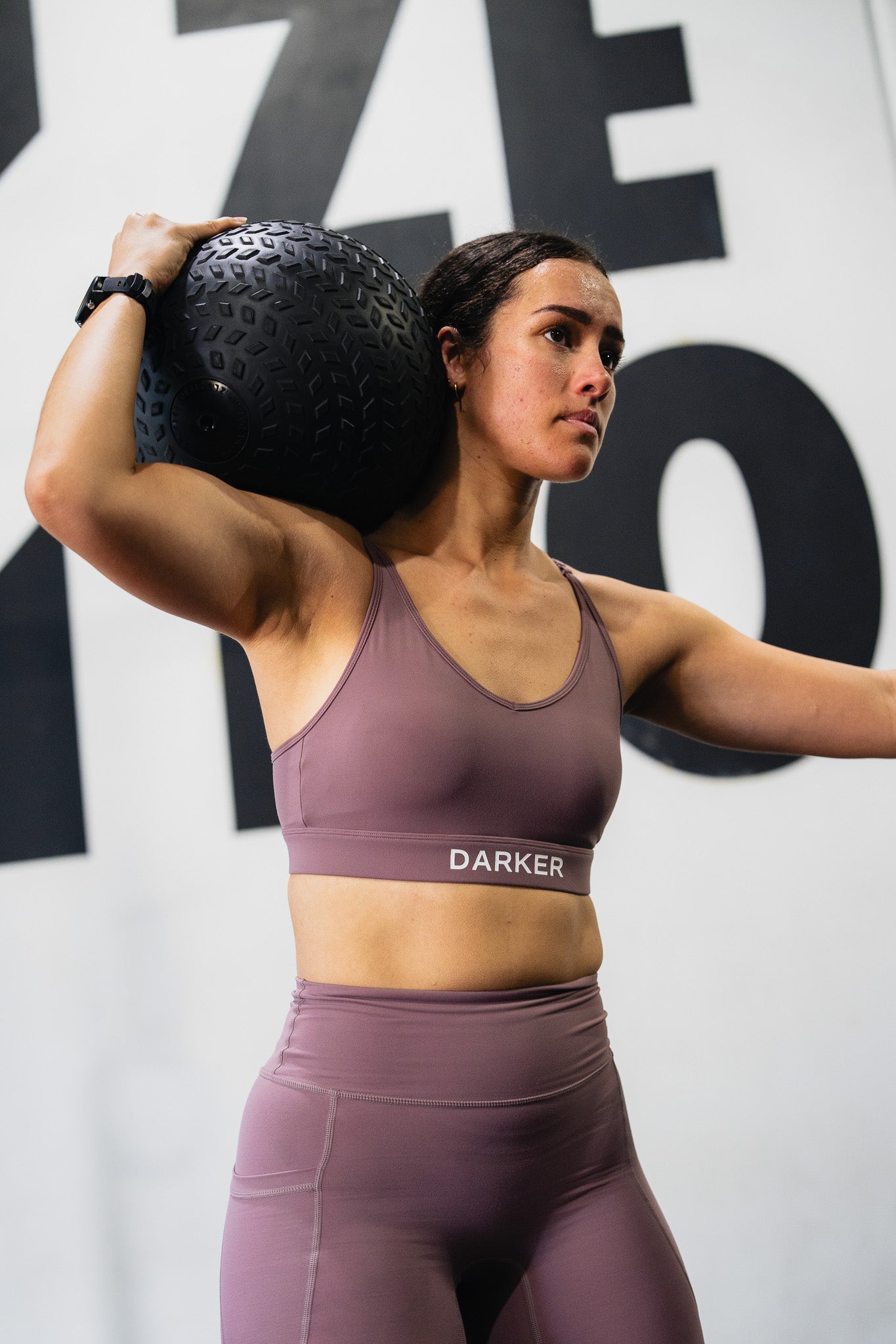 A woman in a gym wearing a mauve-purple sports bra and matching leggings, holding a black medicine ball over her shoulder. The brand name 'DARKER' is printed in white on the sports bra band.