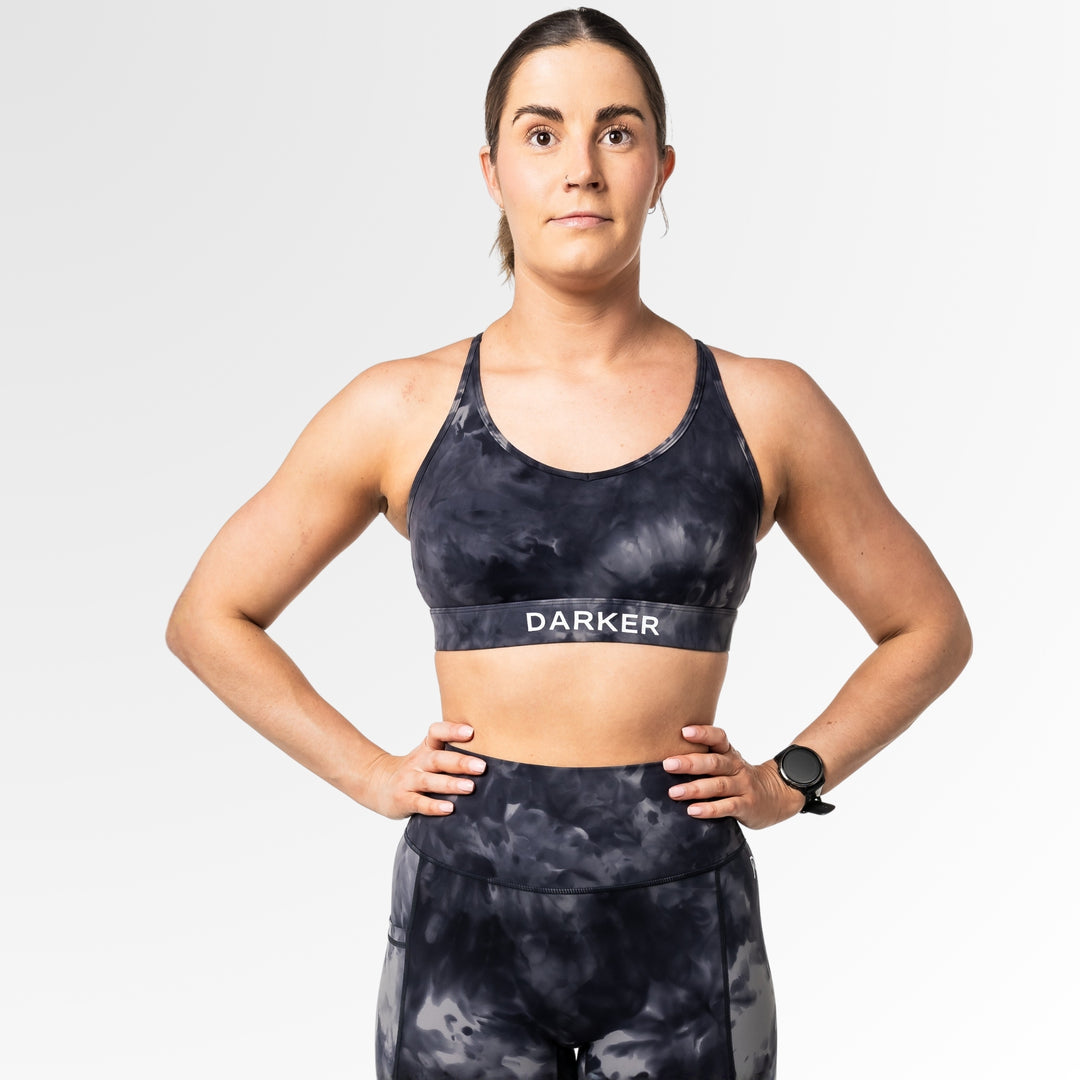 Woman wearing a black tie-dye sports bra and leggings with 'DARKER' branding on a light gray background.