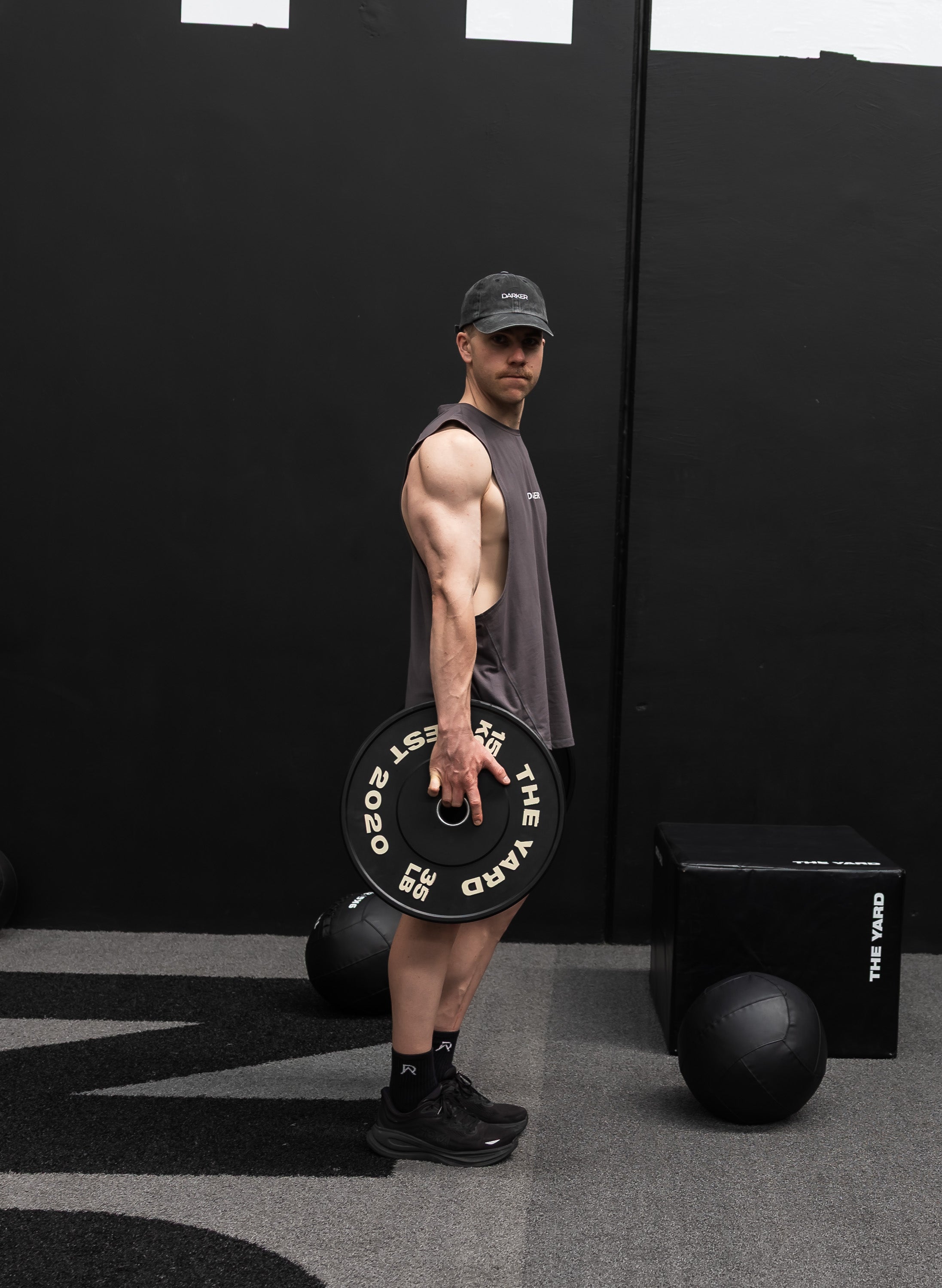 Man holding a barbell plate with gym equipment in the background wearing a black cap